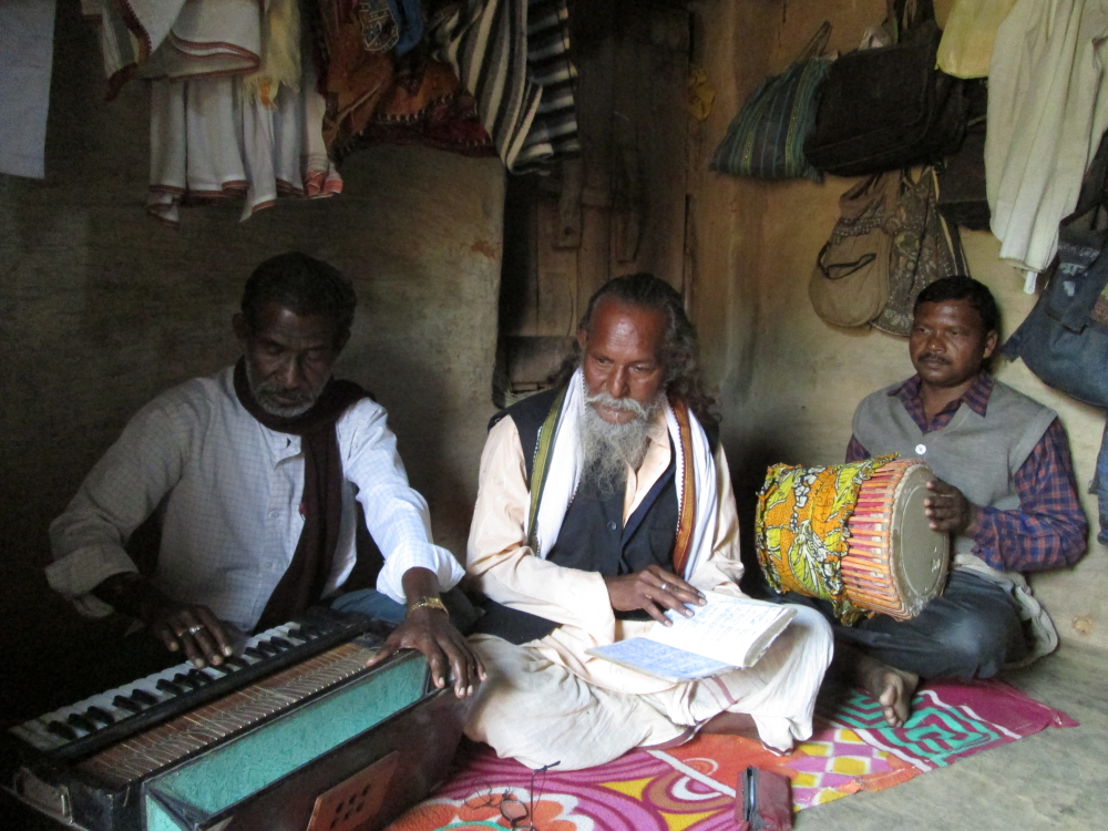 চিত্র ১০. ভিখারিচেলিয়ামা গ্রামের ঝুমুর শিল্পী বিশ্বম্ভর পরামানিক (মাঝের জন)। সত্তরোর্ধ্ব এই মানুষটি ছোট্ট একটি ঘরে থাকেন। তাঁর পেশা বলতে কবিরাজি চিকিৎসা। ঘরের মধ্যেই একপাশে ঝুমুরের বাদ্যসরঞ্জাম, অন্যপাশের তাকে রাখা থাকে কবিরাজি ওষুধ। ইনি মূলত স্বাস্থ্য-সচেতনতা ও পরিবেশ-সচেতনতা মূলক গানই লেখেন। (সৌজন্যে: সোহম দাস)