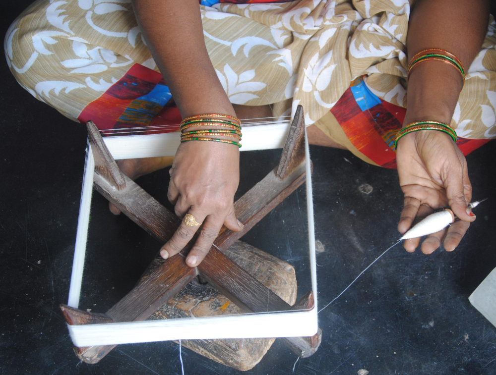 A spinner engaged in making a hank out of handspun yarn. She rotates the frame with her right hand while holding the spindle with her left hand as the yarn from the spindle reels off and winds around the frame. The hank is then taken off the frame, twisted and handed over to the weaver. A handspun hank is 1,000 metre in length (Courtesy: Samyuktha Gorrepati) 