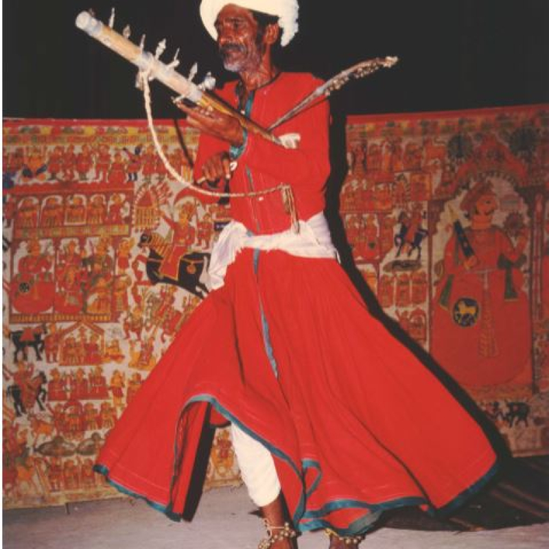 Bhopa Chogo performing the Ravan-hattho stringed instrument in front of the painted backdrop depicting scenes from the Epic of Pabuji